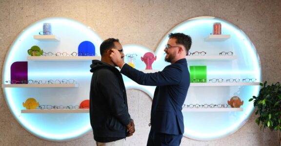 Deux hommes : un ajuste des lunettes à l'autre, étagères colorées en arrière-plan, chaises rouges devant.